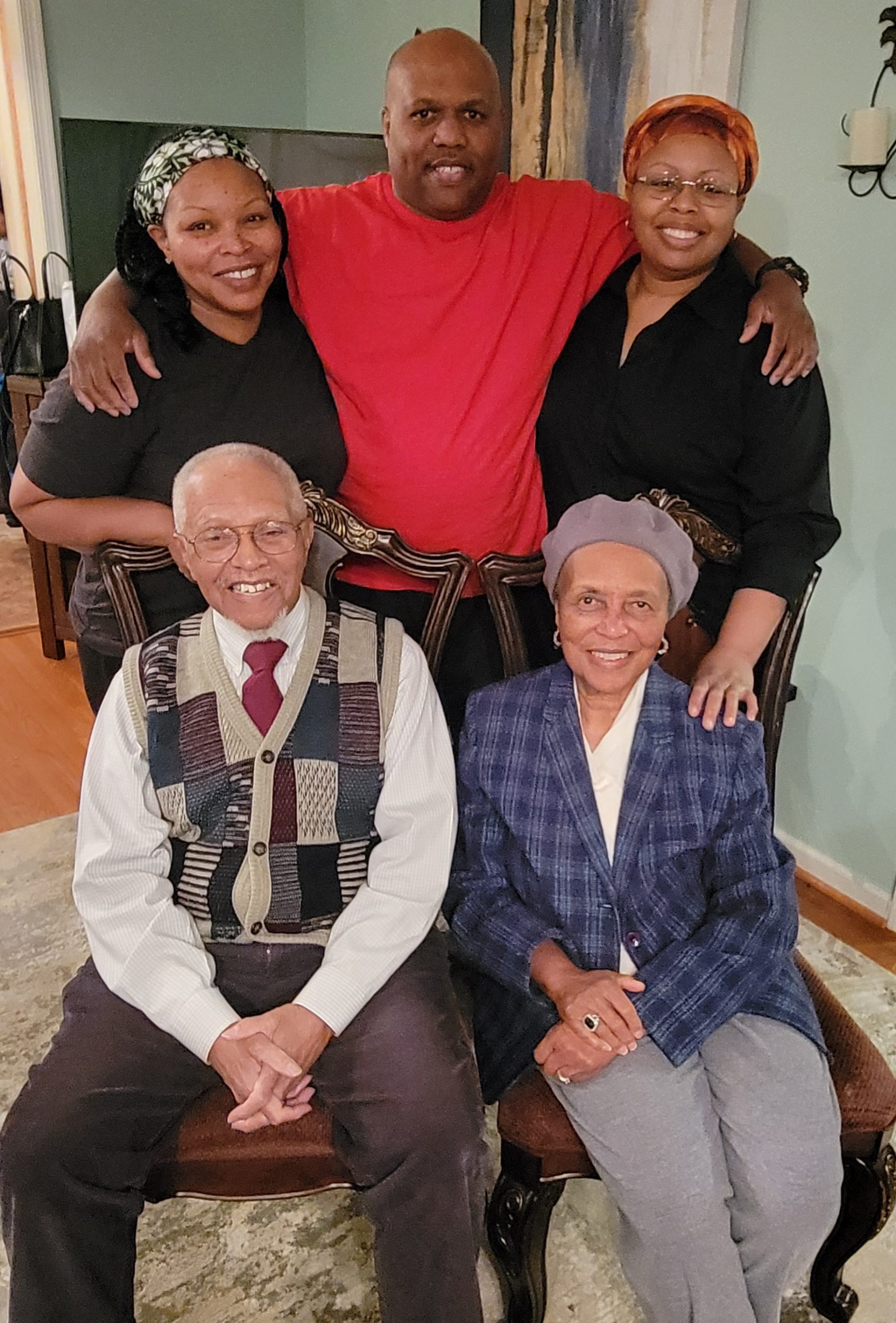 Group of 5 people posing for a picture, three standing up in back, two sitting down in front in chairs